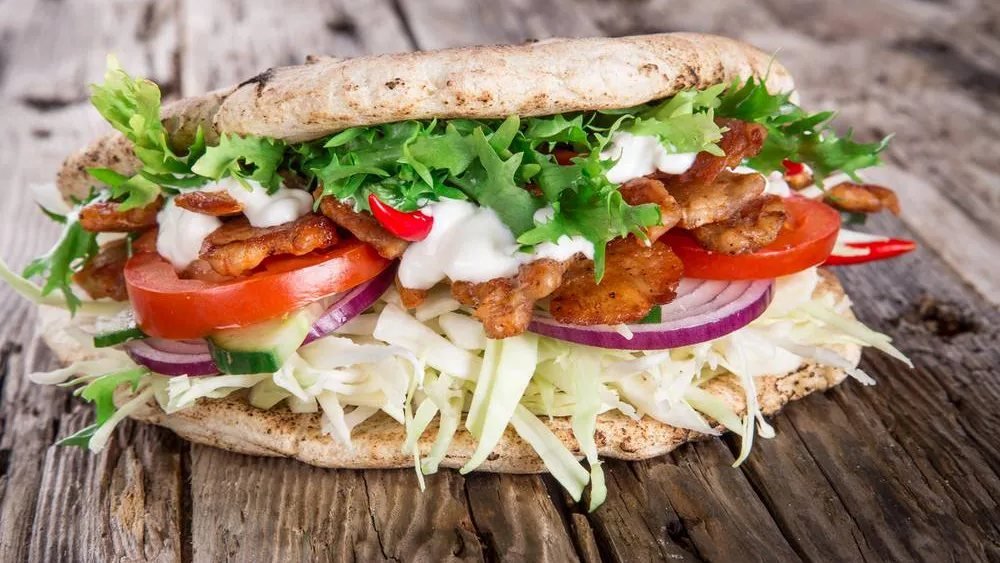 Een pitabroodje met shoarma, ijsbergsla, tomaat, komkommer, rode ui en witte knoflooksaus, op een houten plank.